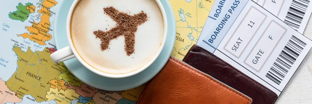 Tasse de café avec une forme d'avion en chocolat sur la mousse, posée sur une carte de l'Europe. À côté, un passeport et une carte d'embarquement avec la mention du siège 11F et de la porte F, illustrant un déplacement professionnel ou un voyage d'affaires.