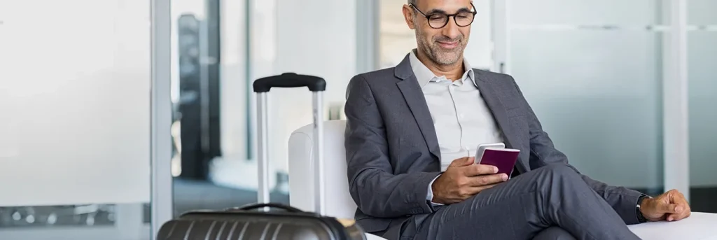 Homme d'affaires assis dans une salle d'attente avec sa valise à côté, utilisant son smartphone et tenant un passeport. L'image montre un professionnel voyageant, symbolisant la gestion efficace des déplacements professionnels offerte par CTA Business Travel.