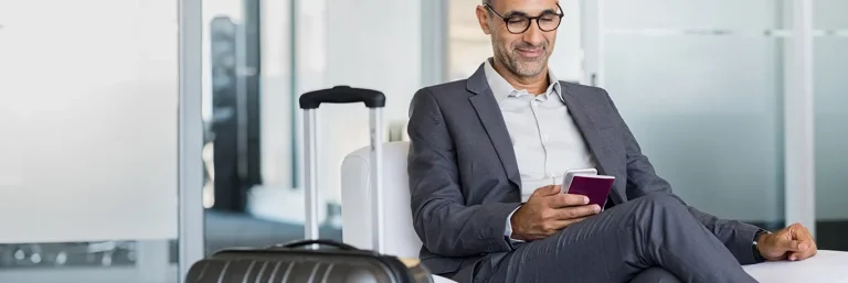 Homme d'affaires assis dans une salle d'attente avec sa valise à côté, utilisant son smartphone et tenant un passeport. L'image montre un professionnel voyageant, symbolisant la gestion efficace des déplacements professionnels offerte par CTA Business Travel.