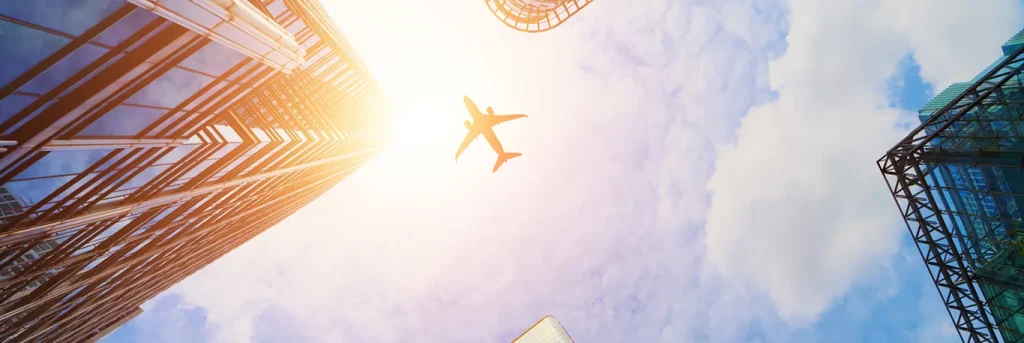 Vue en contre-plongée d'un avion survolant des gratte-ciels modernes en verre sous un ciel bleu et ensoleillé. L'image représente la mobilité et l'efficacité des voyages d'affaires internationaux, illustrant les services de billetterie d'affaires de CTA Business Travel pour des déplacements professionnels optimisés.