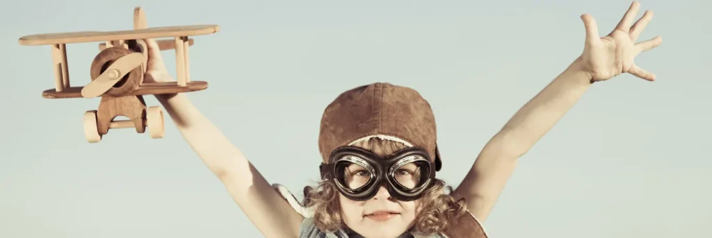 Enfant portant des lunettes d'aviateur et un casque en cuir, tenant un avion en bois avec les bras levés. L'image évoque l'enthousiasme et l'aspiration au voyage, symbolisant un nouveau regard sur les voyages d'affaires, et mettant en avant l'approche innovante de CTA Business Travel pour la gestion des déplacements professionnels.