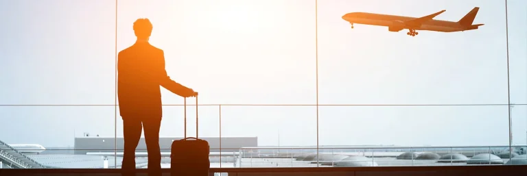 Silhouette d'un homme d'affaires avec une valise à roulettes, regardant un avion décoller à travers une grande fenêtre d'aéroport, baignée par la lumière du coucher du soleil. L'image représente la mobilité et les voyages d'affaires, mettant en avant les services de CTA Business Travel pour faciliter et organiser les déplacements professionnels.