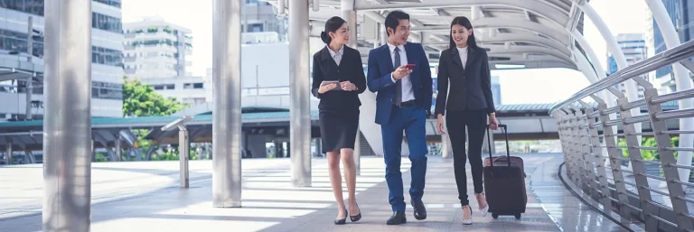 Trois professionnels en costume marchant ensemble dans un environnement urbain moderne, avec des valises et des appareils électroniques. L'image représente la collaboration et la mobilité lors des déplacements professionnels, illustrant les services de CTA Business Travel pour organiser et faciliter les voyages d'affaires pour les cadres et dirigeants.