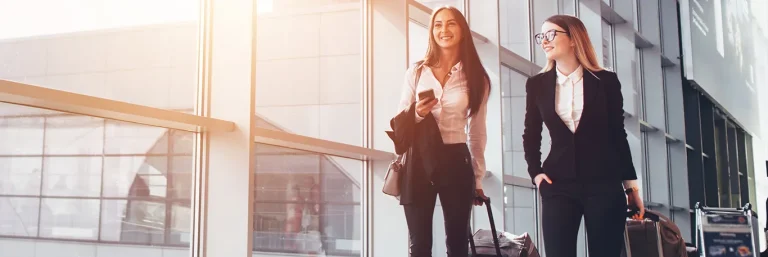 Deux femmes professionnelles souriantes marchant dans un aéroport avec des valises à roulettes et des sacs à main. L'image représente la confiance et la satisfaction lors des voyages d'affaires, soulignant les services de CTA Business Travel pour organiser et faciliter les déplacements professionnels avec efficacité et confort.