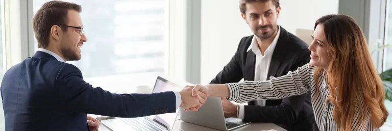 Deux professionnels se serrant la main lors d'une réunion d'affaires, avec un troisième collègue observant, tous assis autour d'une table avec des ordinateurs portables. L'image illustre la collaboration et la réussite des négociations dans le cadre des déplacements professionnels, soulignant les services de CTA Business Travel pour faciliter les rencontres d'affaires efficaces.