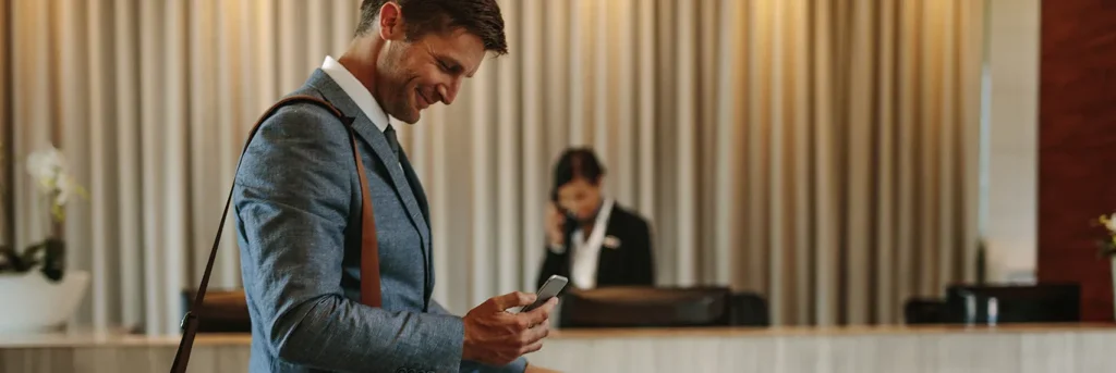 Homme d'affaires souriant, utilisant son smartphone dans le hall d'un hôtel, avec un réceptionniste en arrière-plan. L'image illustre la commodité et le confort des déplacements professionnels, mettant en avant les services de CTA Business Travel pour faciliter les voyages d'affaires et garantir une expérience agréable aux professionnels en déplacement.