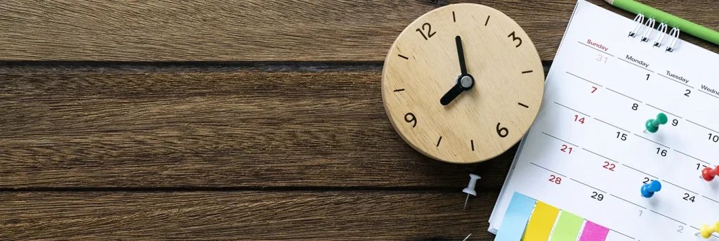 Horloge en bois et calendrier avec des épingles colorées sur un bureau en bois. L'image symbolise la planification et la gestion du temps, essentielles pour organiser des voyages d'affaires efficaces, illustrant les services de CTA Business Travel dans la coordination des déplacements professionnels.