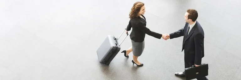 Deux professionnels en costume se serrant la main dans un hall d'aéroport, l'un tirant une valise à roulettes et l'autre portant une mallette. L'image symbolise les voyages d'affaires et les rencontres professionnelles, soulignant l'importance de la préparation et de l'organisation offertes par CTA Business Travel pour des déplacements professionnels réussis.