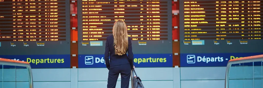 Une femme en tenue professionnelle, de dos, regardant un grand tableau d'affichage des départs dans un aéroport. Le tableau affiche les informations de vol et les destinations. Cette image illustre les voyages d'affaires à l'international, mettant en avant l'organisation et la planification nécessaires pour des déplacements efficaces et bien coordonnés.