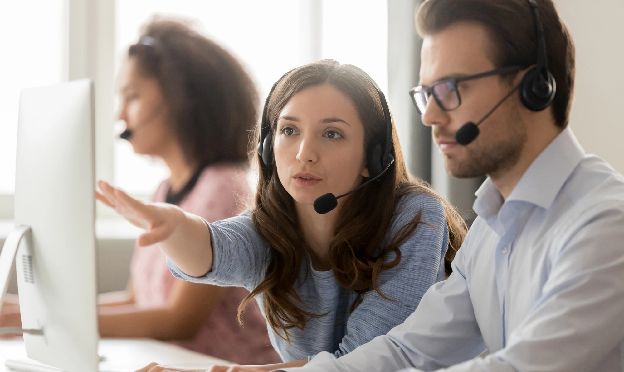 Deux agents de support client, portant des casques avec micro, travaillent sur des ordinateurs. Une femme montre quelque chose à l'écran à son collègue, illustrant la collaboration et l'assistance en temps réel. En arrière-plan, une troisième agente de support est également au travail. Cette image représente le service de gestion de crise et d'assistance immédiate offert par CTA Business Travel pour les voyages d'affaires.