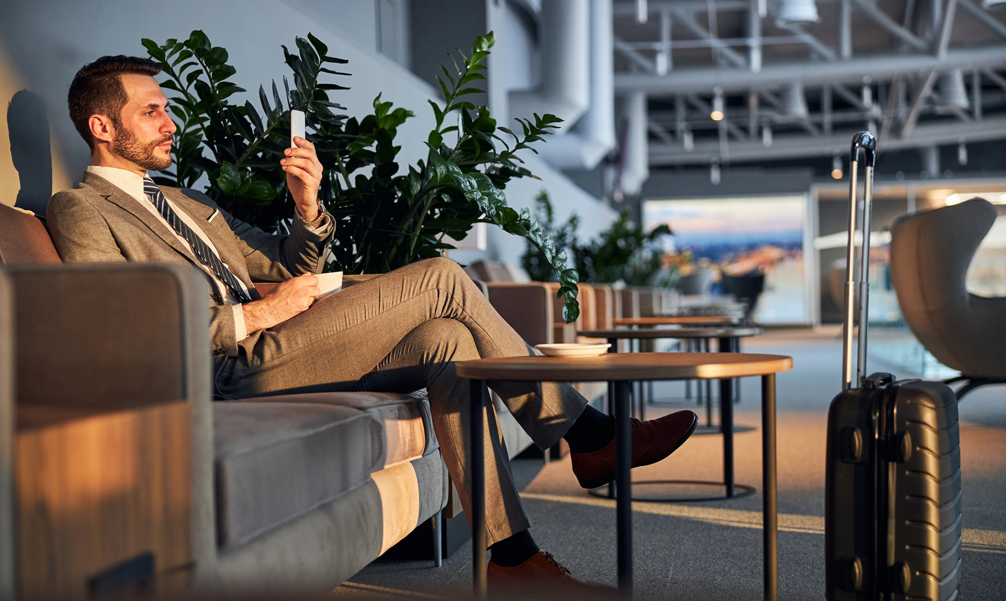 Un homme en costume, assis dans un salon d'aéroport, tenant un smartphone et une tasse de café. À ses côtés, une valise à roulettes. Cette image illustre l'accompagnement et le confort offerts par CTA Business Travel pour les voyages d'affaires, mettant en avant une expérience de voyage fluide et agréable.