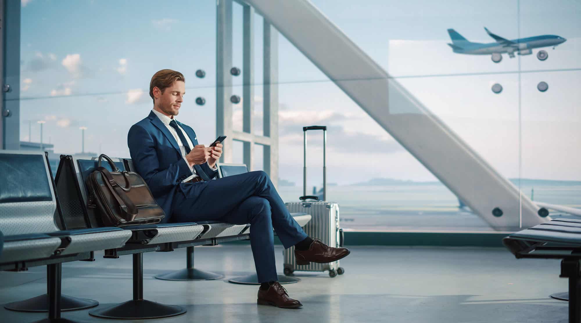 Un homme en costume bleu est assis dans une salle d'attente d'aéroport, regardant son téléphone. À côté de lui, il y a une valise argentée et un sac en cuir marron posé sur le siège adjacent. En arrière-plan, à travers la grande baie vitrée, on peut voir un avion en vol et une vue dégagée sur la piste et le ciel bleu. L'homme semble détendu et concentré, prêt pour son voyage d'affaires. L'ambiance est moderne et professionnelle, reflétant le dynamisme des voyages d'affaires.