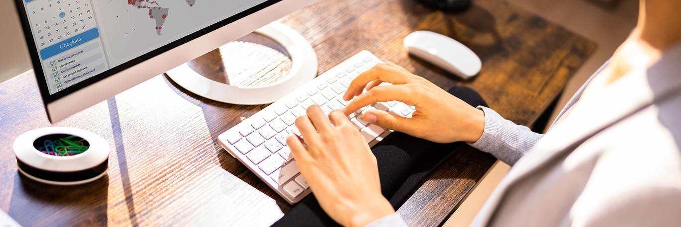 Un professionnel en costume utilise un clavier d'ordinateur sur un bureau en bois. À l'écran, des graphiques, une carte et une checklist sont visibles, suggérant une planification et une gestion des voyages d'affaires. À côté de l'ordinateur, une souris et un objet circulaire décoratif sont présents.