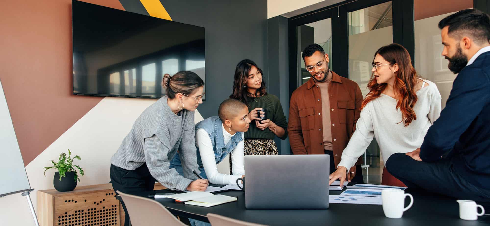 Un groupe de six personnes se tient autour d'une table de réunion dans un bureau moderne et bien éclairé. Ils semblent engagés dans une discussion ou une séance de brainstorming. Une femme à droite, portant un pull blanc, pointe du doigt un document sur la table tandis que les autres écoutent attentivement. Sur la table, on voit un ordinateur portable ouvert, des documents et des tasses de café. L'ambiance est collaborative et dynamique, reflétant le travail d'équipe et la planification stratégique, probablement dans le contexte de la gestion des voyages d'affaires.