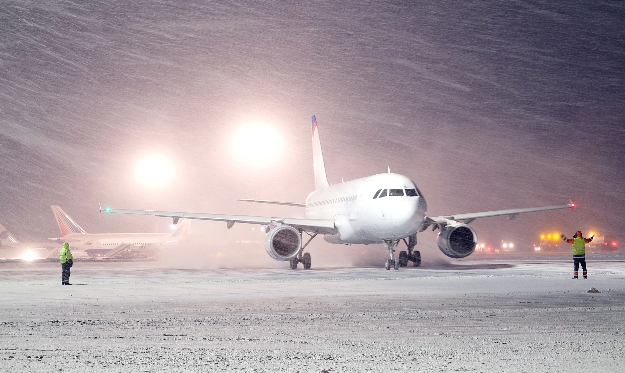 Un avion de ligne sur une piste d'aéroport sous une forte tempête de neige, guidé par deux agents au sol en gilets de sécurité. Des lumières brillantes illuminent la scène, illustrant la gestion des situations de crise et des conditions météorologiques extrêmes pour les voyages d'affaires.