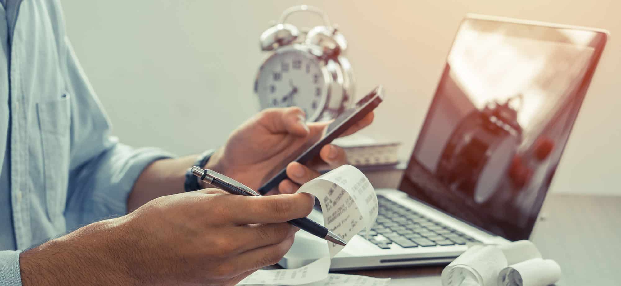 Un homme, vêtu d'une chemise bleue, vérifie des reçus et utilise un smartphone tout en tenant un stylo. Sur la table devant lui, un carnet de notes ouvert, un ordinateur portable, une calculatrice et plusieurs reçus enroulés. Une horloge en métal est visible en arrière-plan. Cette image illustre la gestion des dépenses et la récupération de la TVA pour les voyages d'affaires.