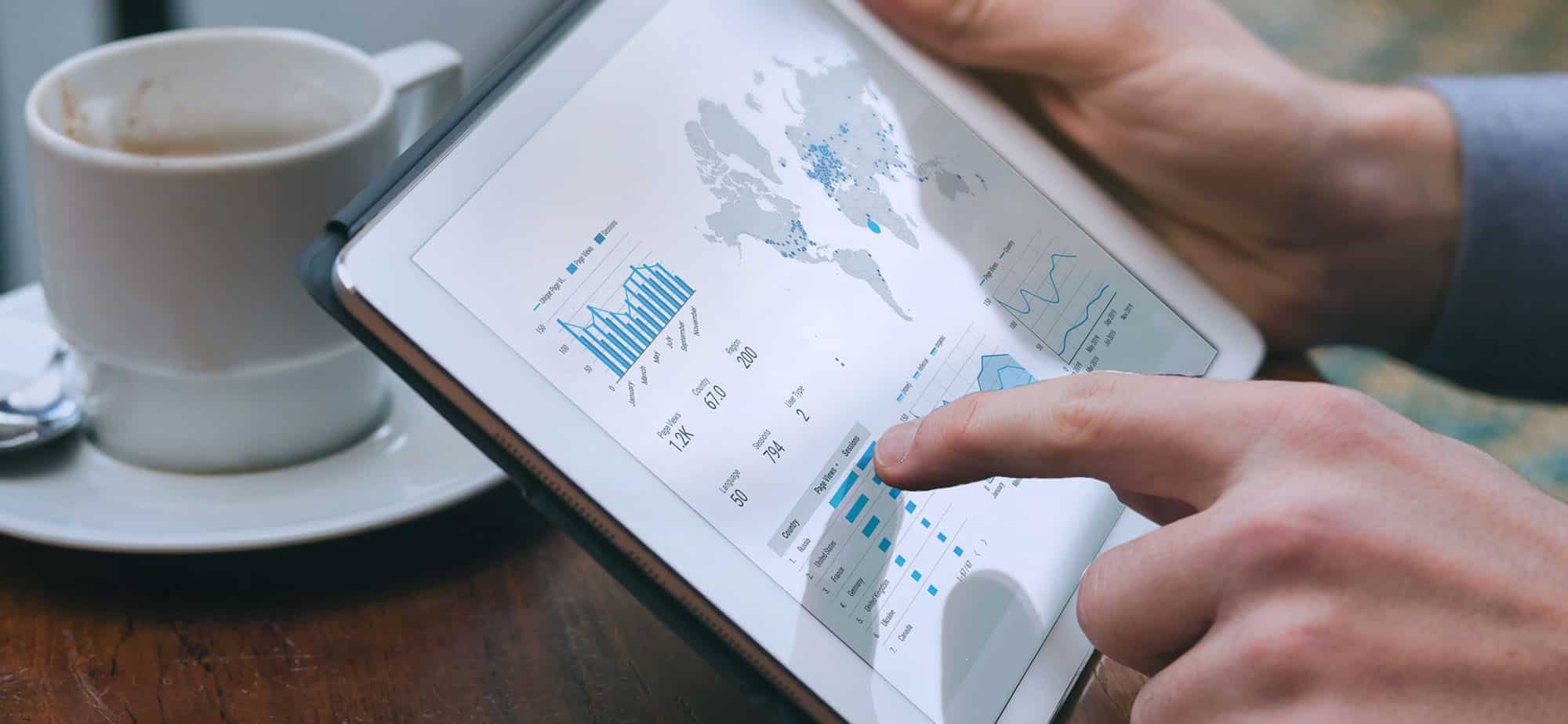 Un gros plan sur les mains d'une personne utilisant une tablette pour analyser des données. L'écran de la tablette affiche des graphiques, des diagrammes et une carte du monde avec des points de données, reflétant des statistiques et des informations de voyage. À côté de la tablette, une tasse de café posée sur une table en bois. Cette image illustre l'utilisation des outils numériques pour le reporting et l'optimisation des déplacements professionnels, permettant une analyse détaillée des voyages d'affaires pour une gestion plus efficace et informée.