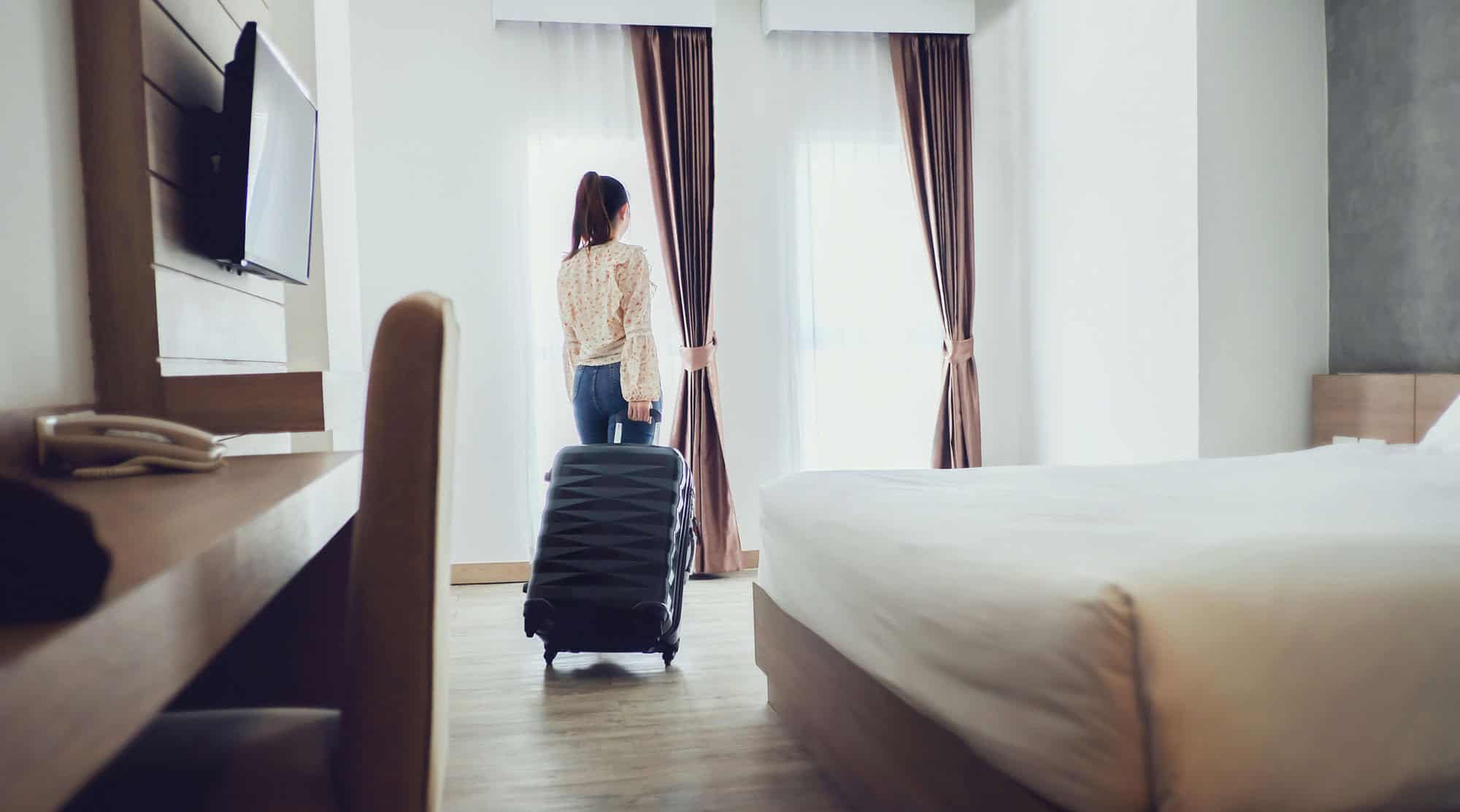 Une femme d'affaire, de dos, se tient dans une chambre d'hôtel moderne avec une valise noire à roulettes. Elle regarde par la fenêtre, encadrée de rideaux marron clair, laissant entrer une lumière douce dans la pièce. La chambre est aménagée avec un grand lit, un bureau en bois avec un téléphone et une télévision montée au mur. L'ambiance est calme et accueillante, parfaite pour un séjour de voyage d'affaires.