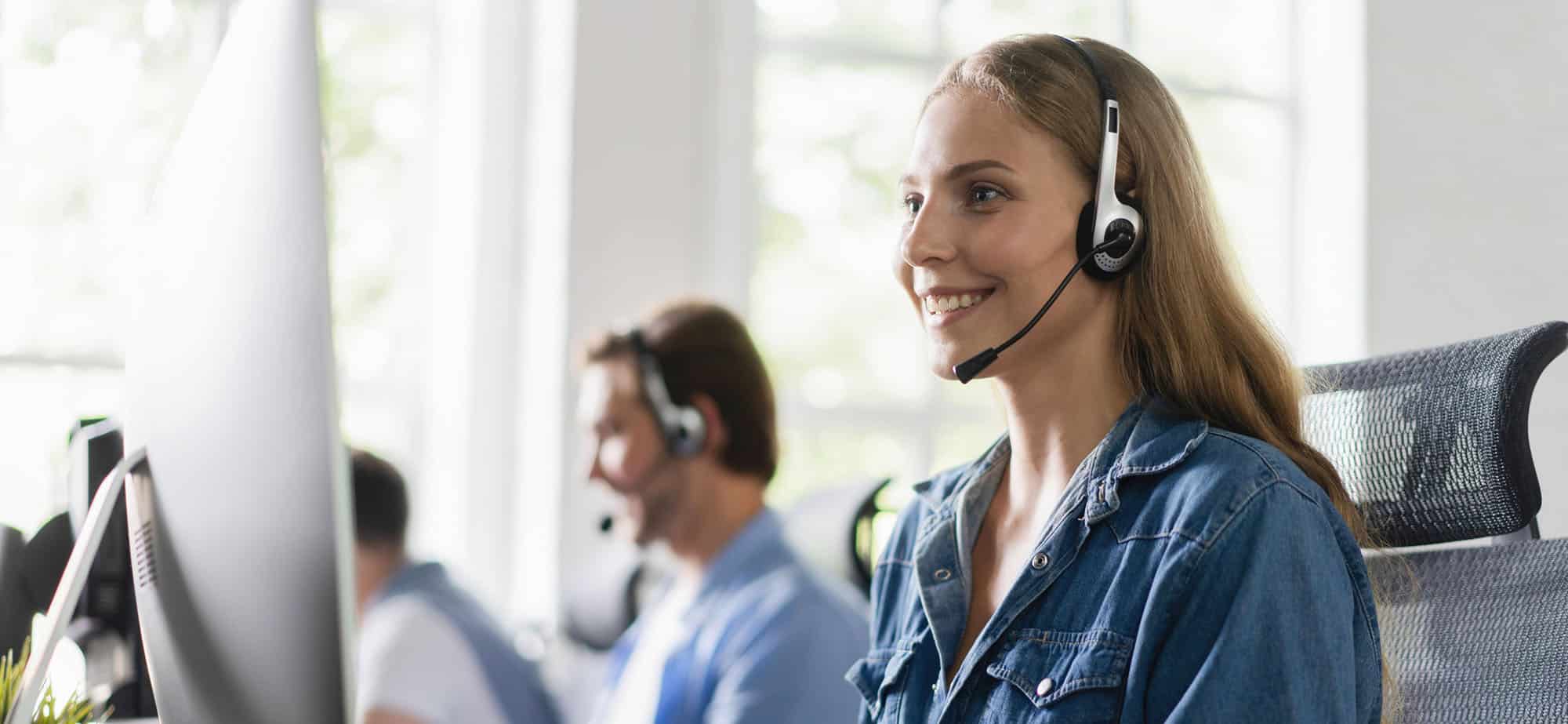 Une jeune femme souriante, portant un casque avec micro, est assise à son bureau et travaille sur un ordinateur. Elle porte une chemise en jean et semble concentrée tout en interagissant de manière professionnelle. En arrière-plan, plusieurs collègues, également équipés de casques, travaillent à leurs postes dans un espace de bureau lumineux et moderne. Une tasse de café est posée à côté de l'ordinateur de la femme, suggérant un environnement de travail dynamique et collaboratif, dédié à la gestion des comptes et au service client dans le domaine des voyages d'affaires.