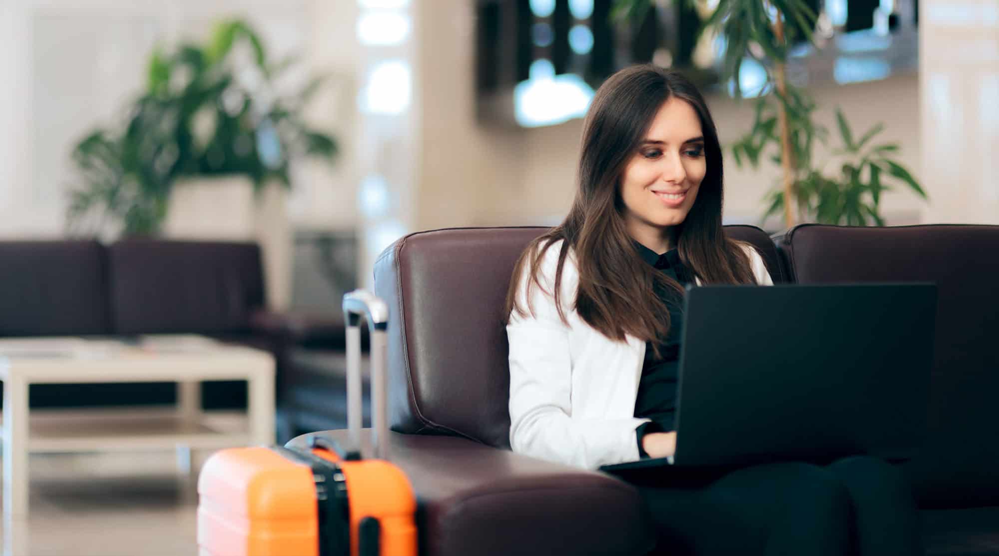 Une femme est assise sur un fauteuil en cuir dans une salle d'attente moderne, travaillant sur son ordinateur portable. Elle sourit et semble concentrée. À côté d'elle, une valise orange est posée, indiquant qu'elle est en déplacement. L'arrière-plan est lumineux et flou, avec des plantes et des meubles modernes, créant une ambiance accueillante et professionnelle. Cette image illustre bien le confort et la connectivité nécessaires pour les voyages d'affaires.