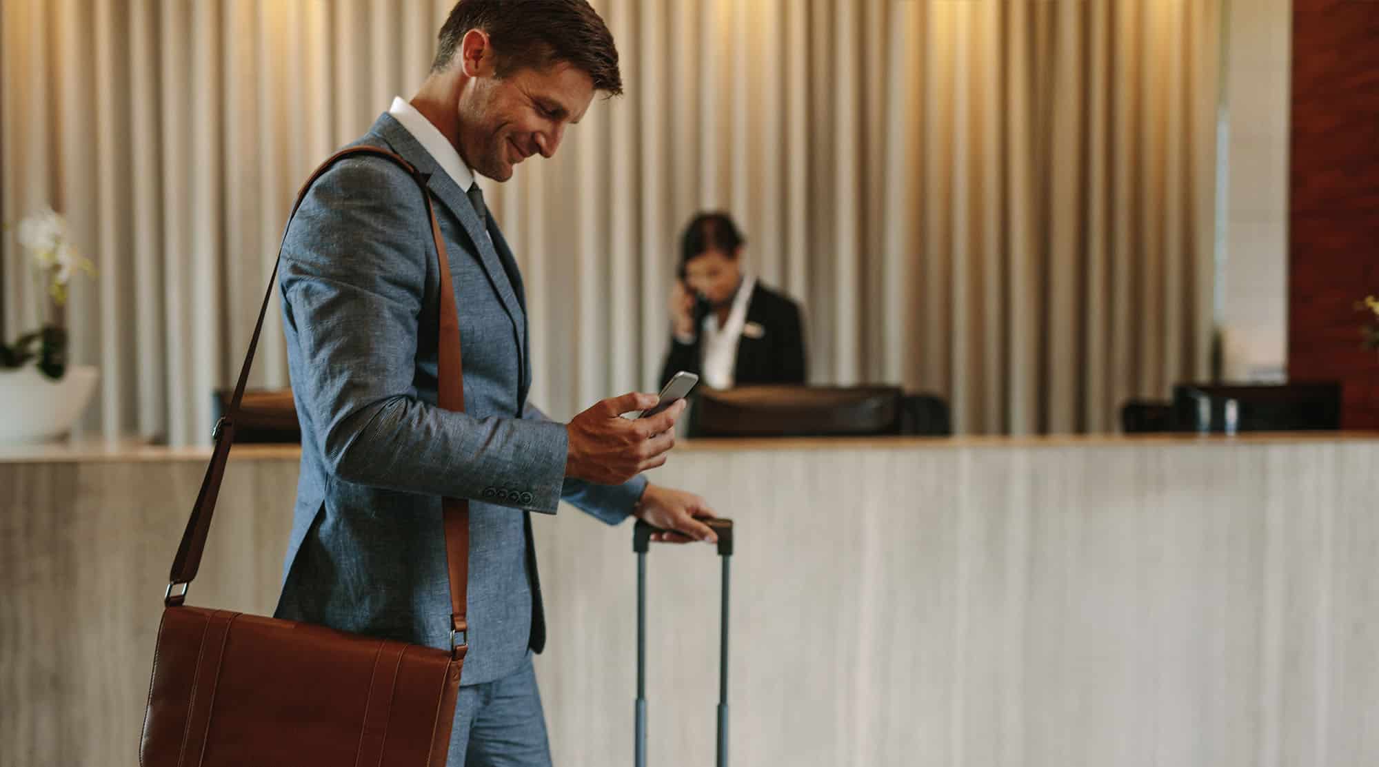 Un homme en costume, portant une sacoche en cuir marron en bandoulière, se tient dans le hall d'un hôtel avec une valise à roulettes. Il regarde son téléphone avec un sourire, suggérant qu'il vérifie des informations ou envoie un message. En arrière-plan, le comptoir de réception de l'hôtel est visible avec une employée parlant au téléphone. La scène dégage une atmosphère professionnelle et accueillante, typique des déplacements d'affaires bien organisés.