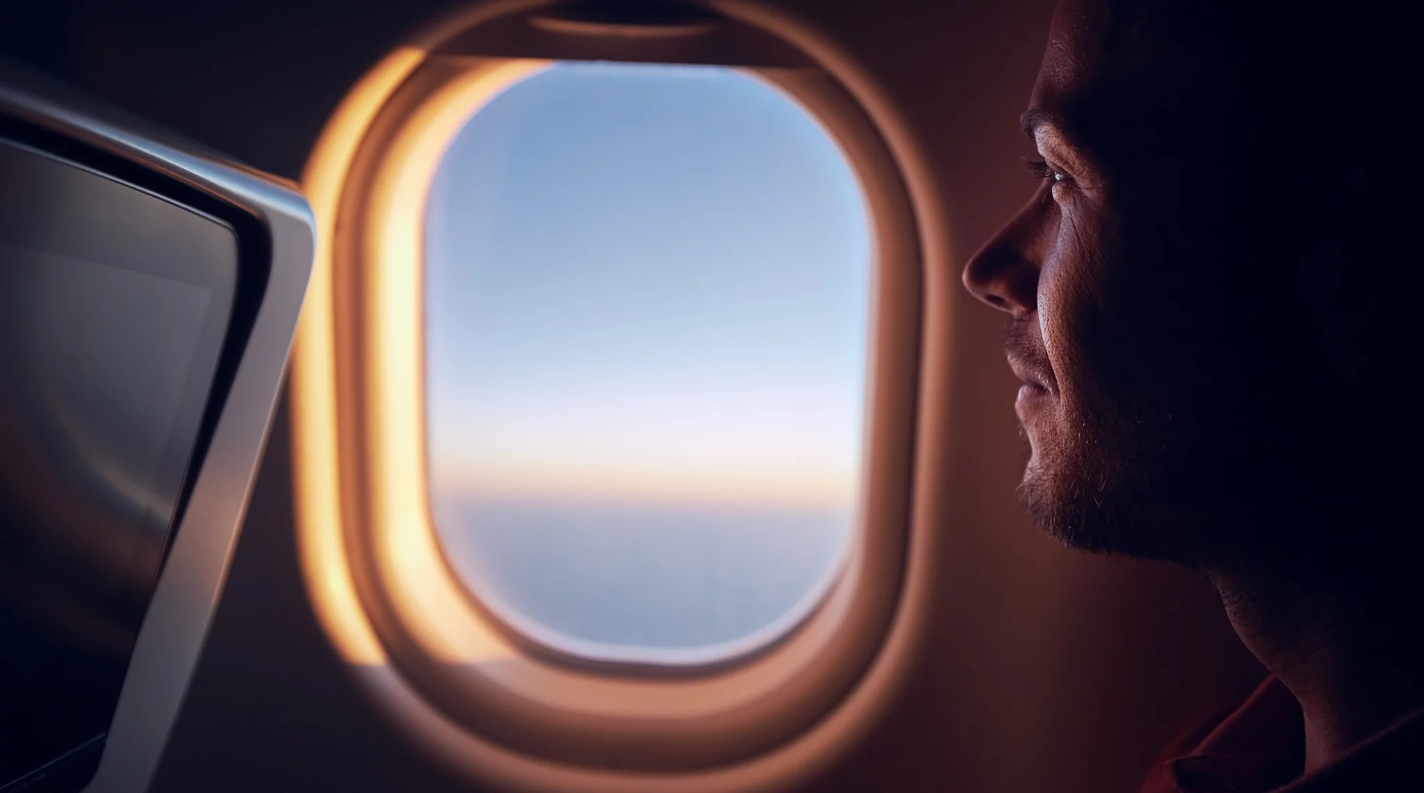 Un homme est assis dans un avion, regardant par le hublot. Son visage est éclairé par la lumière douce du coucher ou du lever du soleil, créant une ambiance chaleureuse et contemplative. À côté de lui, un écran de télévision intégré au siège de devant est partiellement visible. À travers le hublot, on peut voir le ciel dégagé et l'horizon lointain.