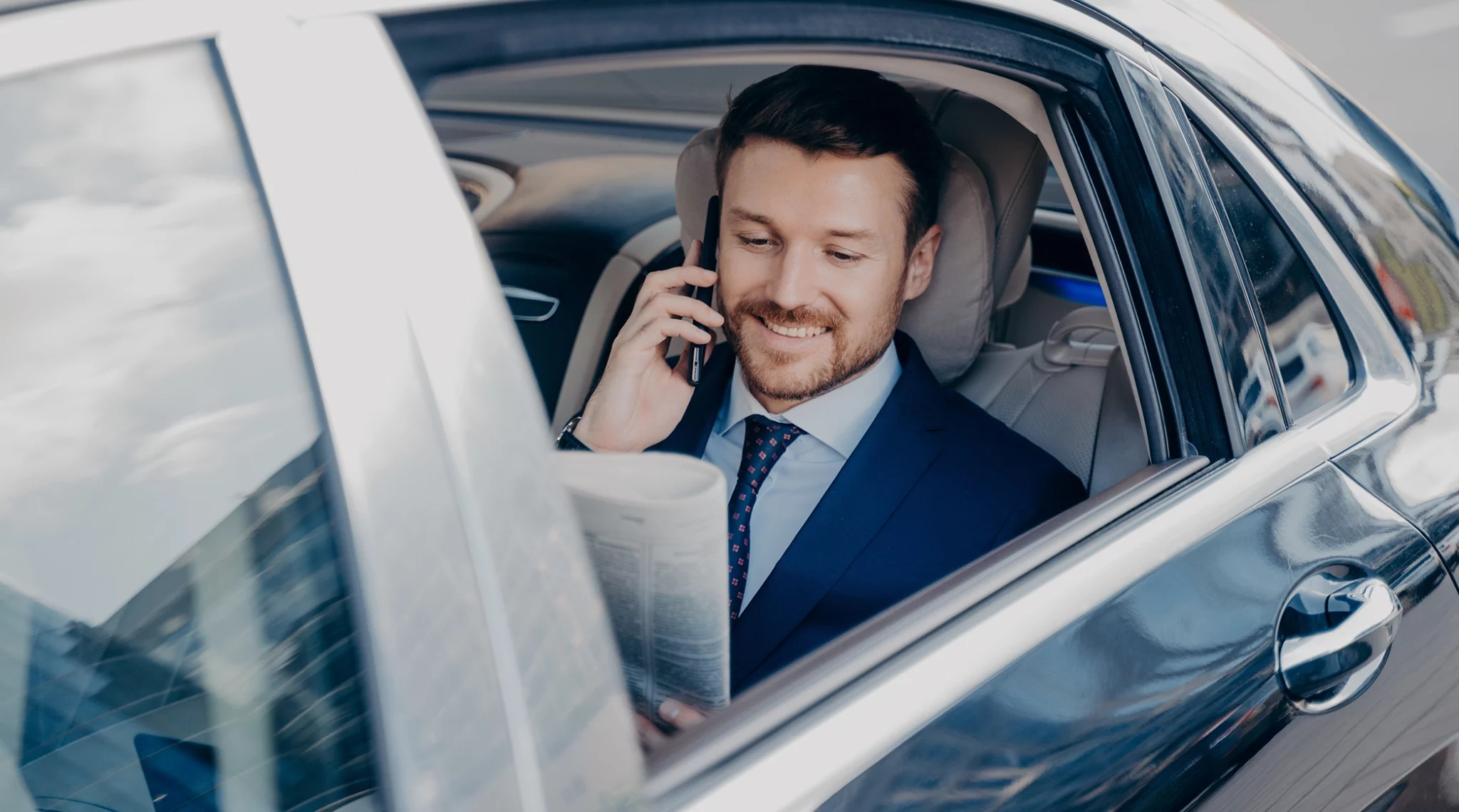 Un homme en costume, assis à l'arrière d'une voiture, est en train de téléphoner tout en lisant un journal. Il sourit et semble détendu. La scène se déroule en plein jour et les reflets des immeubles environnants sont visibles sur les vitres de la voiture, suggérant un environnement urbain. Le confort de la voiture et l'attitude de l'homme suggèrent qu'il est en déplacement professionnel.